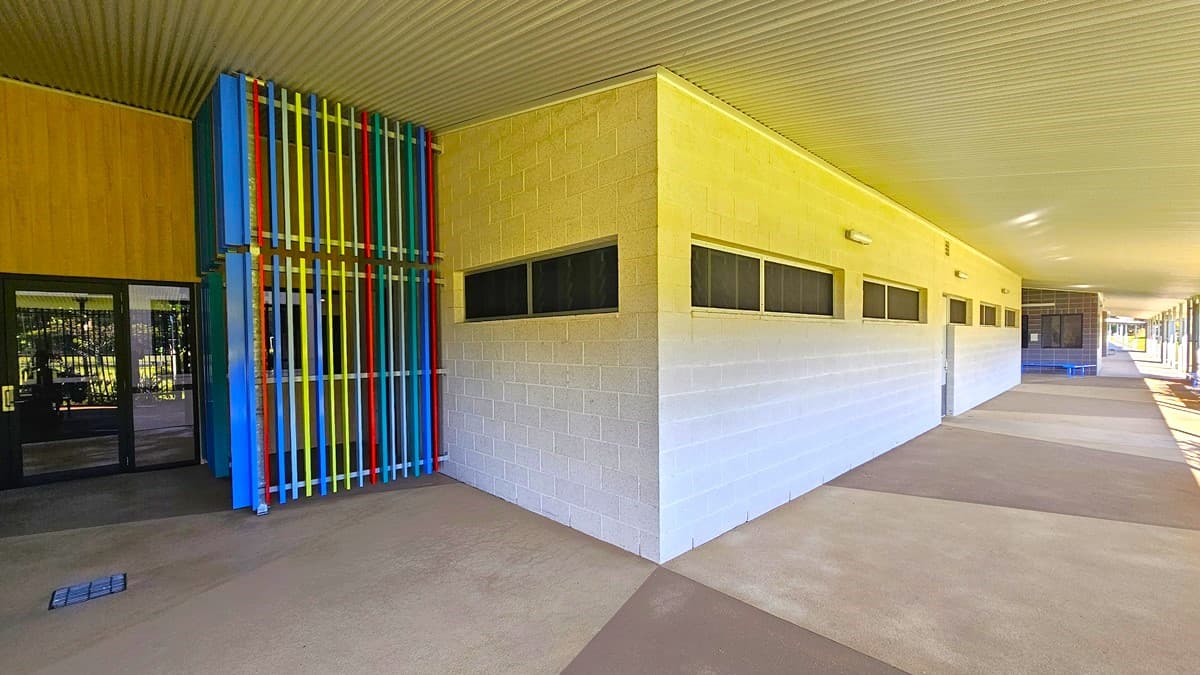 On the left, there is a building entrance with glass doors and a colourful structure made of vertical blue, yellow, red, and green bars. In the middle, there is a long beige wall with small rectangular windows near the top. On the right, the wall continues with more windows and a blue door in the distance. The floor is a mix of beige and brown concrete, and the ceiling is a white corrugated metal roof.