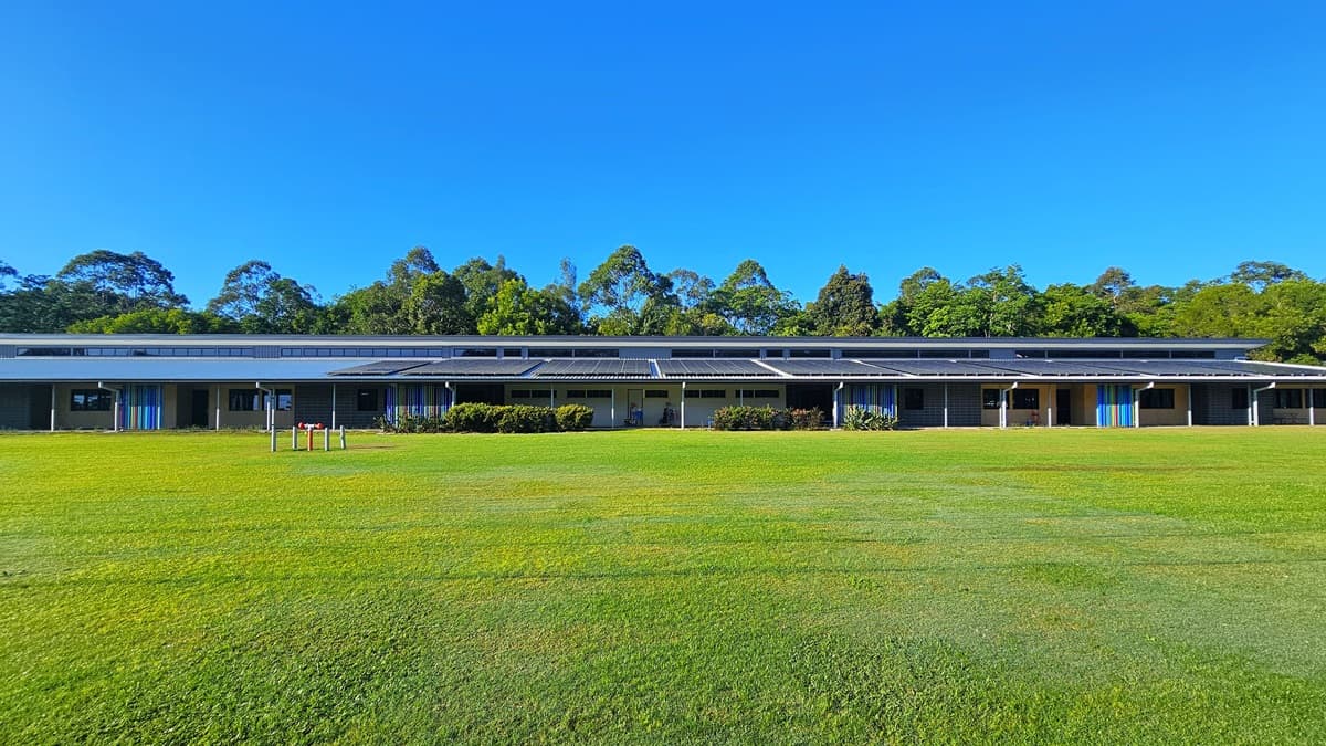 On the foreground, there is a grassy field with a few small wooden structures. In the middle, there is a long, single-storey building with a dark roof, featuring multiple windows and doors, and a row of bushes in front. On the right, the building continues with more windows and doors, and a blue door is visible. The floor is a well-maintained grassy field, and the ceiling is a dark, flat roof. In the background, there are tall trees behind the building.