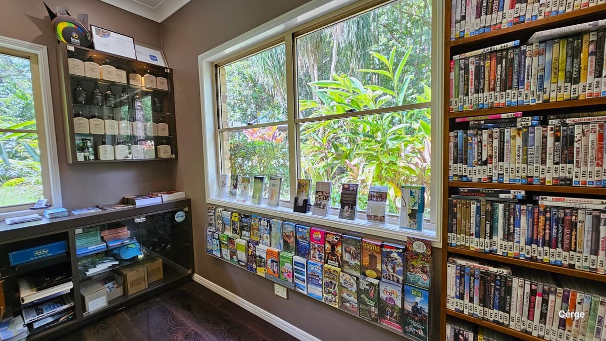Upon arrival and turning right from the entrance at Narrows Escape Rainforest Retreat, you'll encounter a room featuring shelves filled with DVDs for rent, tourism information, retail items, and games available for borrowing, all neatly stacked. A large glass window on the right provides a view of the surroundings. In the corner, another shelf displays awards information. The floor is made of dark timber, complementing the vintage brown wall.
