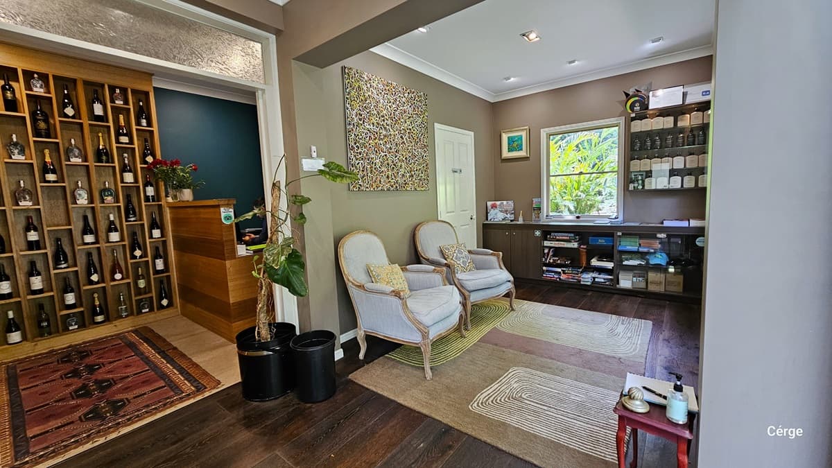Upon entering Narrows Escape Rainforest Retreat's reception, a wall displays various wines near the desk, with a carpet in front. Two cushioned white chairs are against the right wall. A cabinet holds tourism info, retail items, and awards details. A small tea table is in the corner. The ceiling has a white cove design, and the wall is almond and vintage brown. An abstract art painting is on the wall. The floor is timber, partially covered with an intricate carpet.