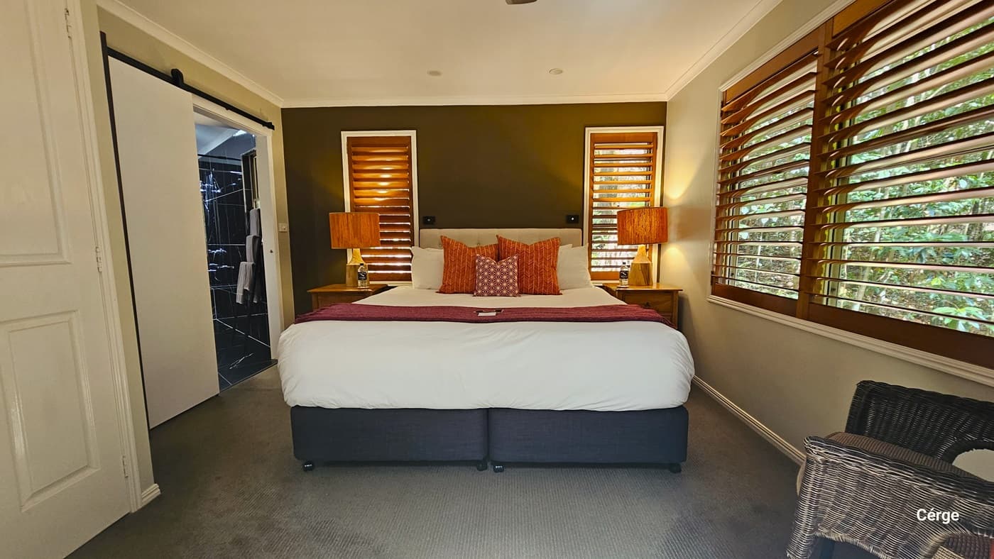 A queen-size bed occupies the center, adorned with tasteful decor. On either side, table lamp shades in coordinating colours with the bed linens illuminate the space. Three windows provide natural light, one large window to the right and two smaller ones behind the bed headrest, each adorned with brown window blinds. To the left, a glimpse of the accessible bathroom is visible, while to the right, a rattan-designed chair is positioned. The ceiling and walls are painted in a pristine white hue.