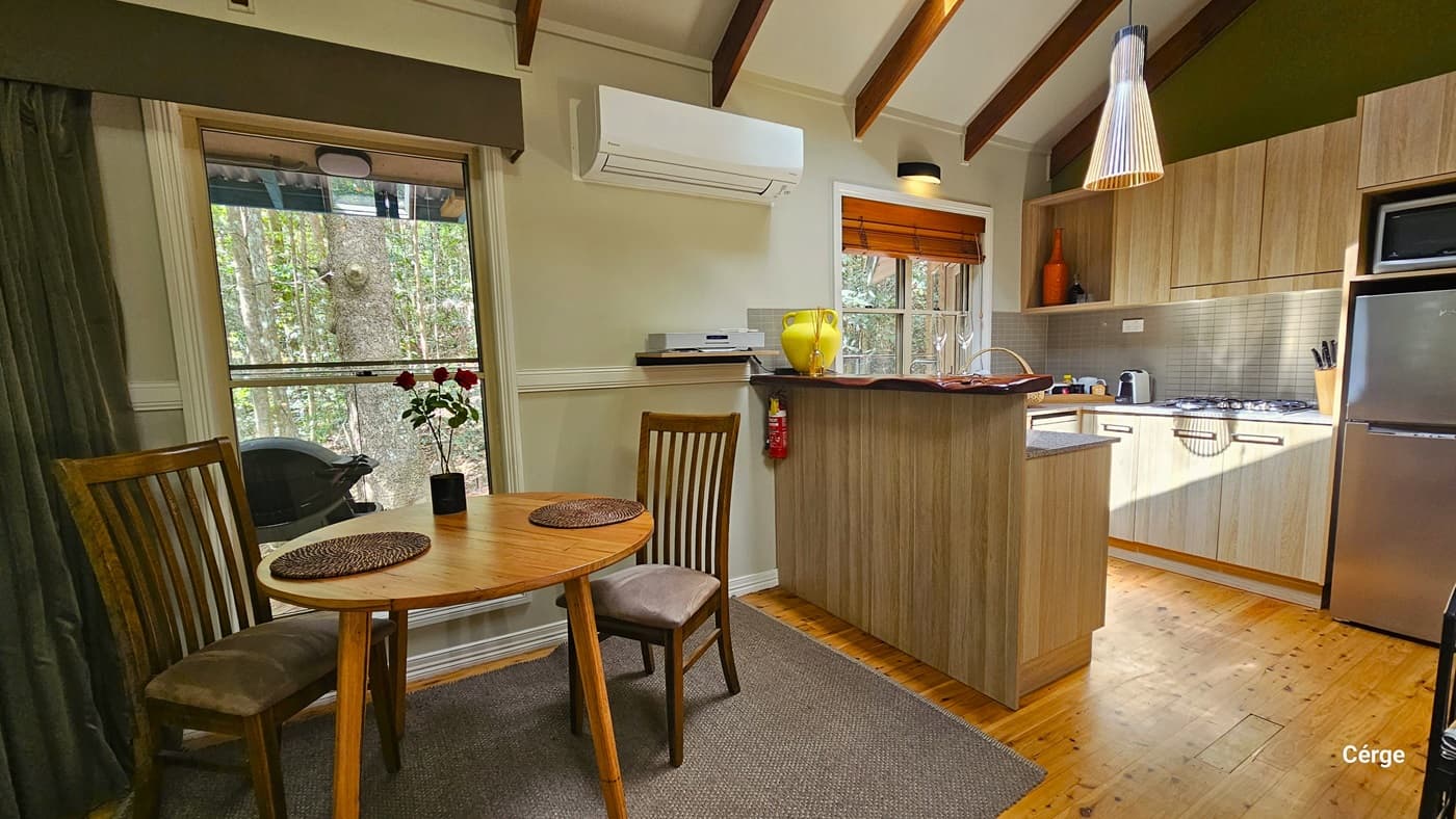 In the Dove Cottage kitchen, timber flooring supports a round table with two chairs, adorned with a flower vase and table mats. A glass window offers a view of the rainforest, complemented by an olive curtain. To the right, a tiled kitchen sink, fridge, stove, and accessories are arranged. There is also a mini kitchen island near the table. Wall-mounted air conditioning and lighting fixtures illuminate the space, while the ceiling features a wooden plank design with a hanging light fixture.