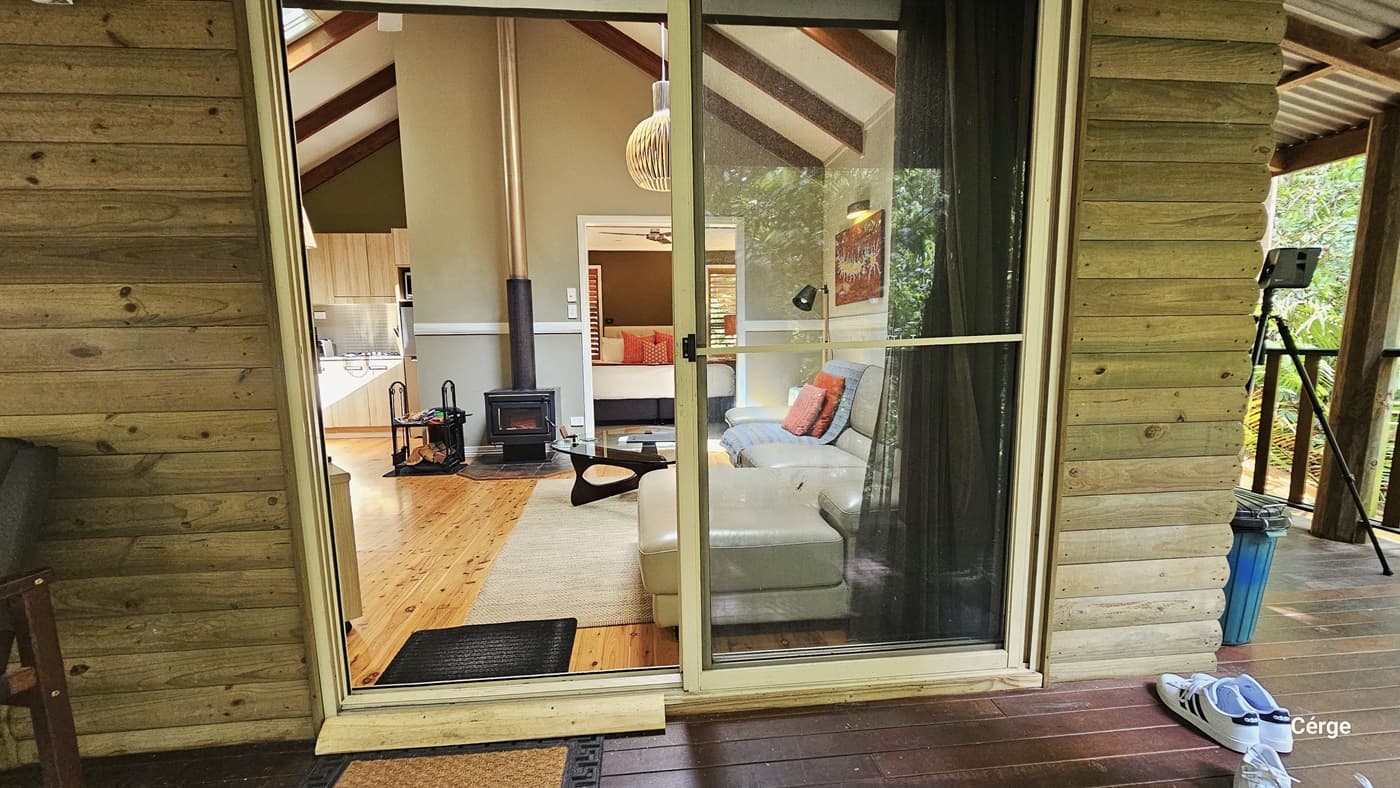 In this view, a glass sliding door serves as the entrance to the cottage. Both the interior and exterior floors are made of timber. Inside, a cream leather couch adorned with pillows and a glass center table are present. On the right wall, there is a framed decoration and a hanging light fixture from the ceiling. A mini shelf displays magazines and chopped wood for decoration. Outside, on the verandah, a cushioned chair is placed, with floor mats positioned before the door entrance.
