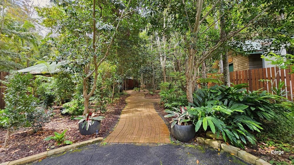 Approaching Kookaburra Cottage, to the right of the road is a shaded carport where a red car is parked. A mini trolley stands nearby. On the left, a plant box bears a signage reminder. Further to the left, there is a row of shaded carports. Tall trees, shrubs, plants, and grass surround the area.