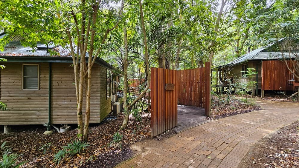 Accessing Kookaburra Cottage, a building with a wooden plank design wall and a green corrugated roof stands along the path. A separate path, bordered by a wooden fence with a sign, is present. The ground area is uneven, surrounded by trees, plants, and dry leaves covering the ground. To the right, another building with a wooden plank wall is visible.