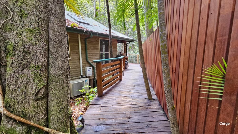 Accessing Kookaburra Cottage, the path is constructed with wooden planks, and a corresponding fence surrounds the building. On the left, a large tree is visible. The building's wall is adorned with air conditioning units and pipes. Pine trees are present in the surroundings