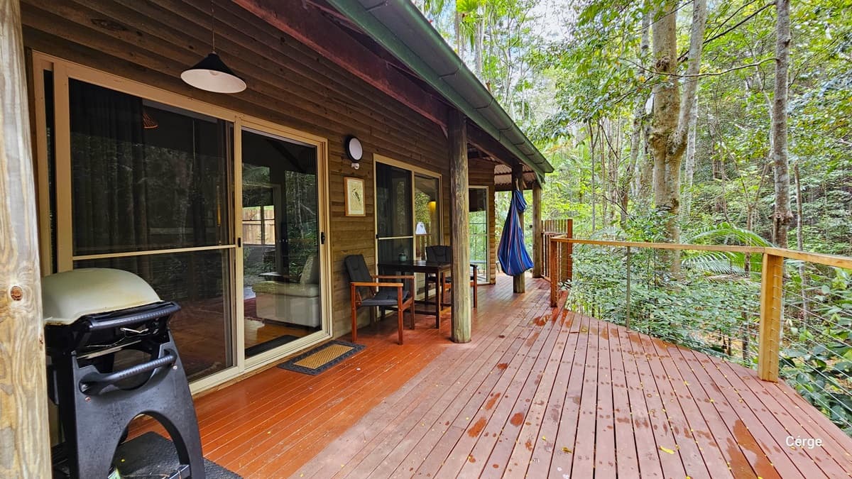 This is the verandah serving as the entrance to the cottage. The building features a wooden plank design, and the floor is constructed with timber. A metal railing secures the area. In this view, large trees are visible on the right. On the left, a sliding door is present with a floor mat placed before it. A cushioned chair and table are positioned to the right of the door. To the left of the door, a barbecue grill is located. Lighting fixtures and wooden frame decorations are also visible.