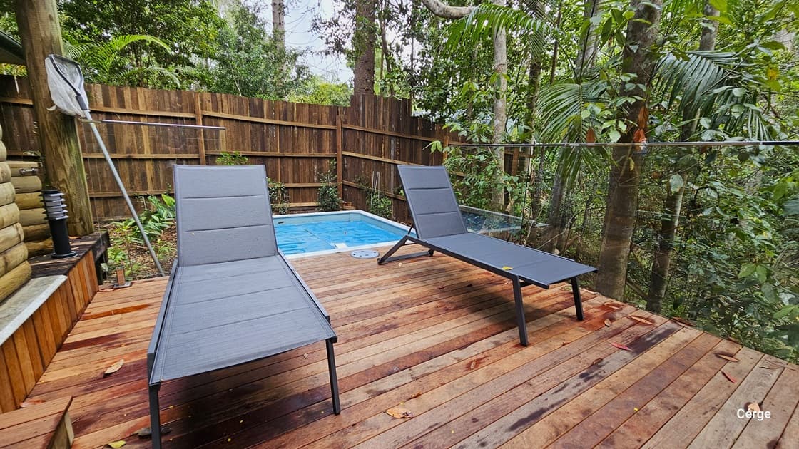 Accessing Kookaburra Cottage. The verandah features a small plunge pool in the end corner, accompanied by two black loungers. The area is secured by a steel fence, with a wooden plank fence providing additional privacy around the pool. The floor is made of timber, and on the left, there is a small lamp post. A leaf skimmer is leaning against a large post. Trees and vines surround the area.