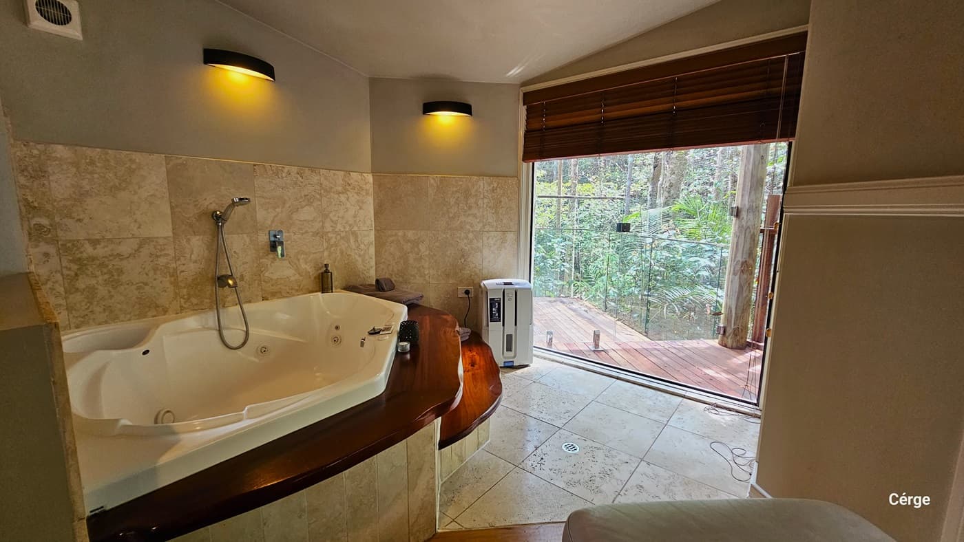 Inside the bathroom, there is a white bathtub fixed against the wall, accompanied by accessible two-step wooden stairs. The bathroom area is painted cream, and the wall features marble design tiles. Lighting fixtures are affixed to the left wall, and a portable heater is placed in the corner. From this vantage point, a view of the forest green outside is also visible.