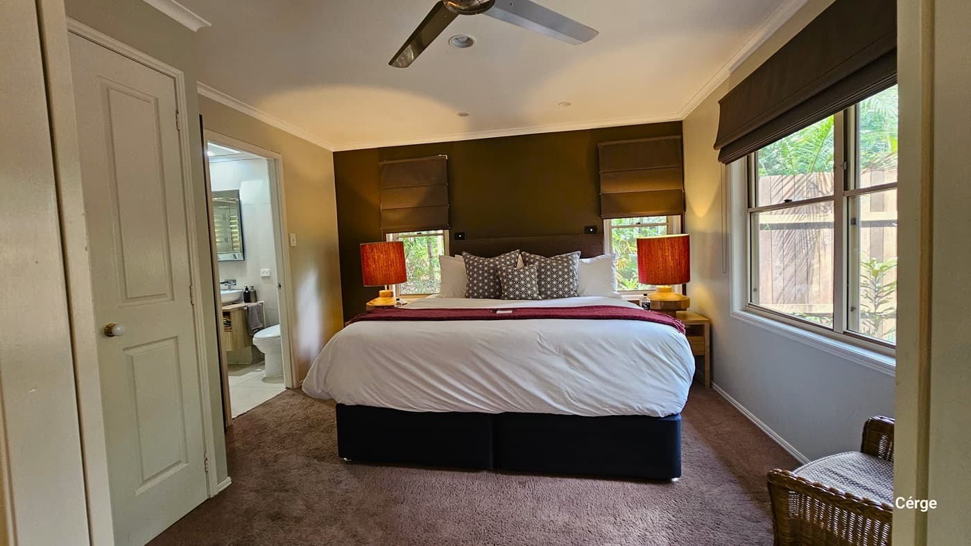 In the middle of the room, a large bed is neatly adorned with pillows and bed linens. Wooden-colored lamps on two side tables flank the bed. To the right, there's a sliding window, and two smaller windows above the bed, all equipped with thick brown blinds. In the left corner, a cushioned rattan chair is placed, and a glimpse of the bathroom is visible. The floor is covered with a brown furry carpet, and a ceiling fan is installed on the white ceiling.