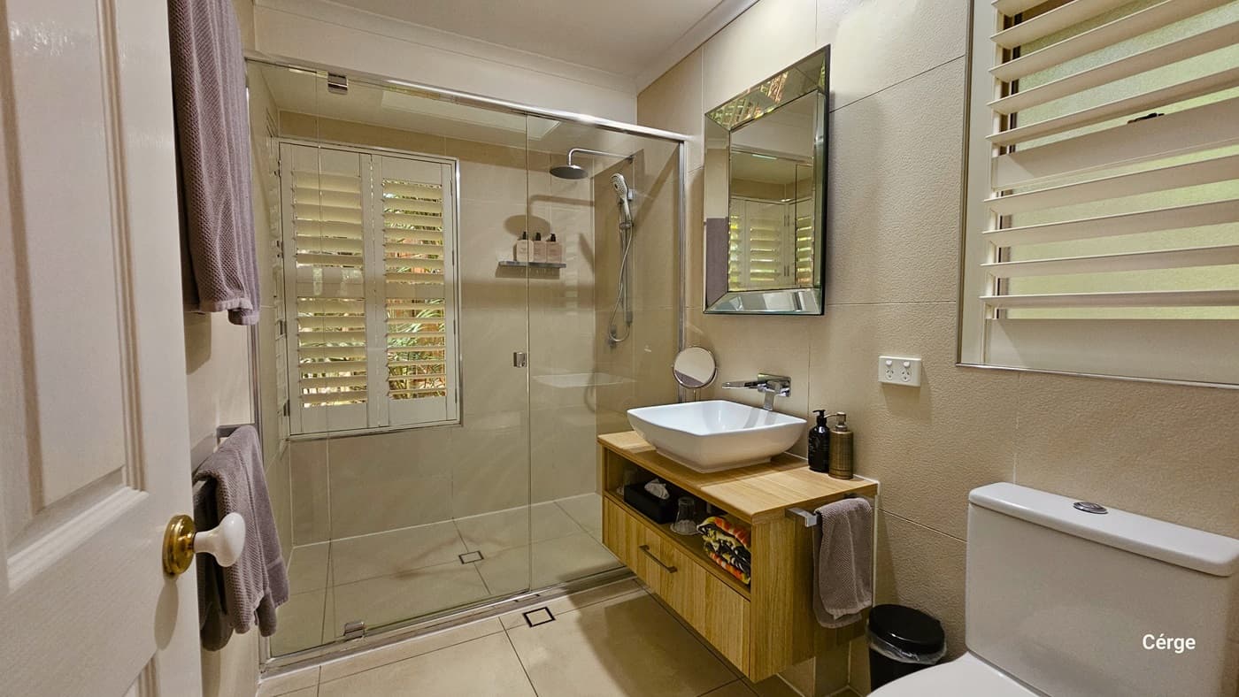In the bathroom, the toilet is situated near the door on the right, accompanied by a black rubbish bin and a wash basin. A bathroom organizer is positioned underneath the wash basin, holding bathroom essentials. On the wall, a mirror is affixed, along with a small round mirror. Next to it, a glass door encloses the shower area, and windows with blinds are installed on both sides. On the left, a rack holding towels is in place. The bathroom is tiled in cream, and the walls and ceiling are white.