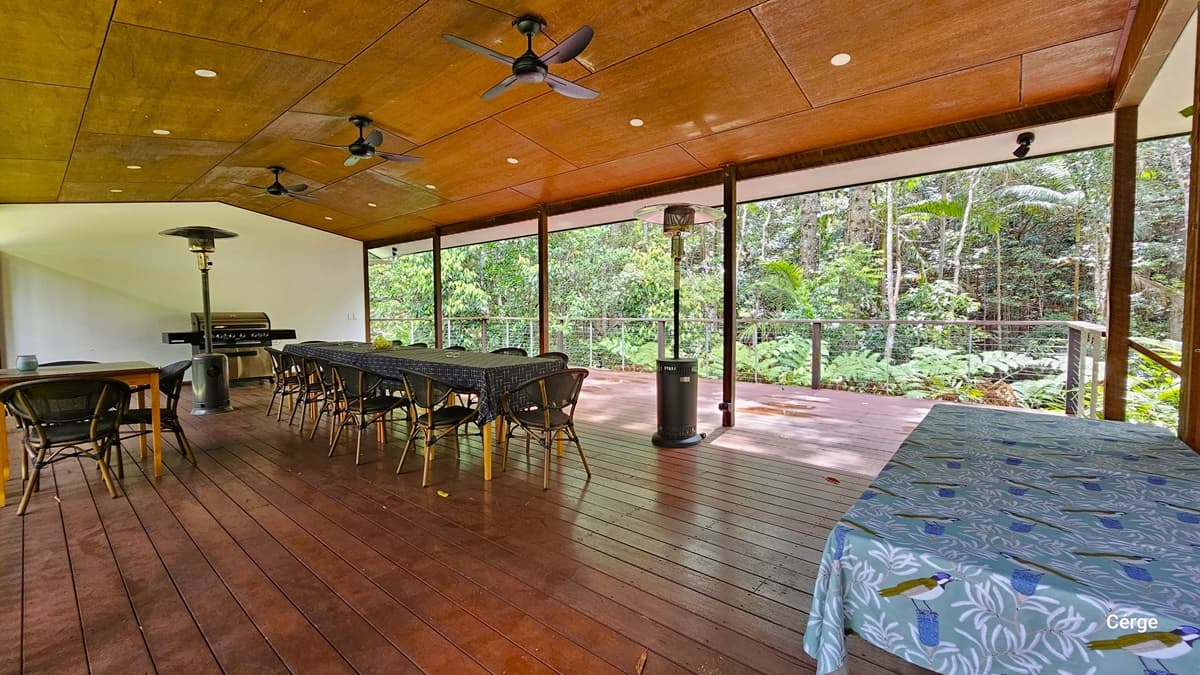 The Rainforest Deck is a spacious area with brown timber plank flooring and a slightly pitched wooden brown roof. Three ceiling fans and a portable heater are installed. Tables and chairs are arranged throughout, including one with a blue tablecloth featuring birds and foliage design. Towards the back, an open verandah secured by metal railings provides views of the surrounding rainforest.
