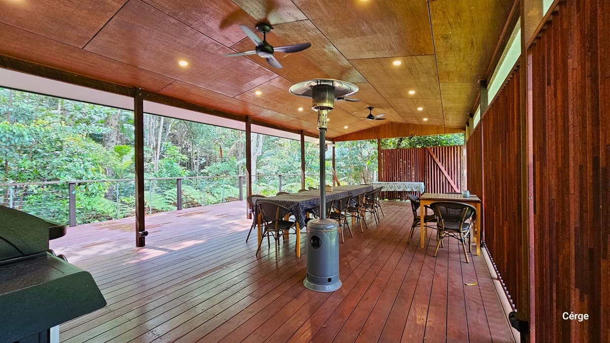 The Rainforest Deck is a spacious area with brown timber plank flooring and a slightly pitched wooden brown roof. Three ceiling fans and a portable heater provide comfort. Tables and chairs are arranged to the sides and the middle. A wall with a wooden plank design extends to the right towards the back. On the left, there's an open verandah secured by metal railings offering views of the surrounding rainforest.