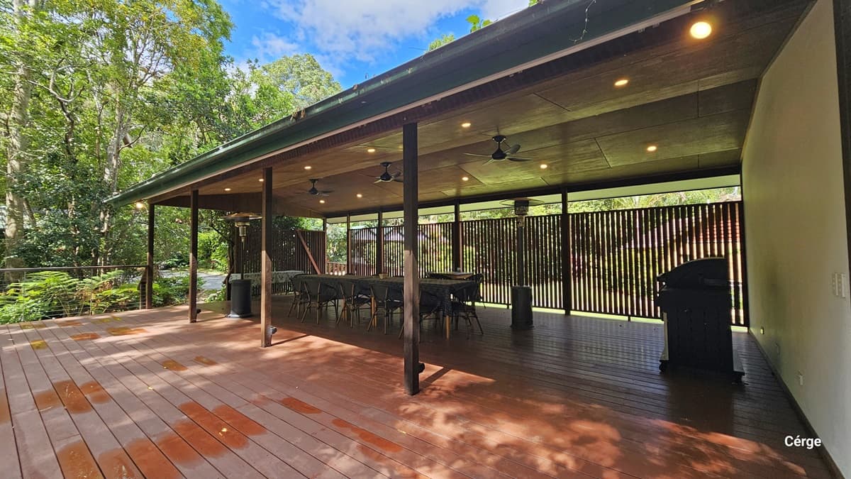 The expansive Rainforest Deck boasts brown timber plank flooring, a gently sloped wooden roof in brown, adorned with three ceiling fans, and a portable heater. Tables and chairs are meticulously arranged along the sides and in the central area. On the right, a wall featuring a wooden plank design extends towards the rear. To the left, there is an open verandah secured with metal railings that provide captivating views of the surrounding rainforest.
