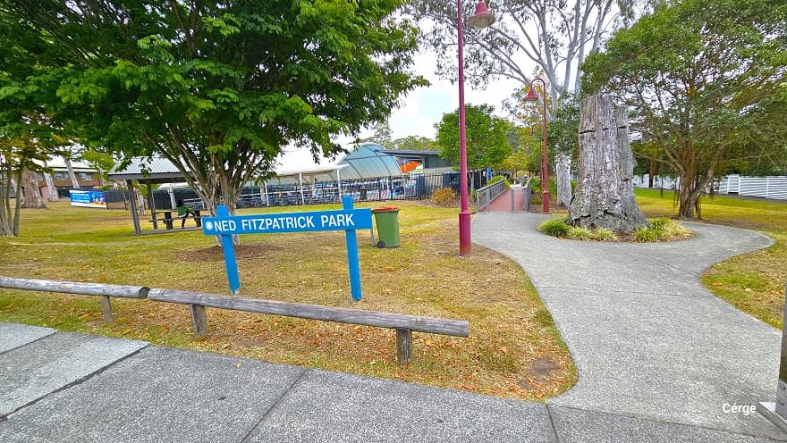 Arrival at Nerang Aquatic Centre from Cotton Street with a red brick road. This road is sloping downward with a number of designated car park spaces marked by white and yellow lines. On the right are residential buildings with similar designs and are enclosed by a white fence with trees. Tall trees with a large canopy cover surround the facility. The red bell-shaped lamp posts and a large white dome-shaped structure are evident from this position.