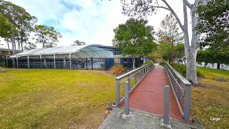 There are two grey rectangular bollards at the start of the red bridge with grey guardrails with strings of wire underneath. There are also red bell-shaped lamp posts next to this bridge. A black fence separates the outdoor amenities from the park.