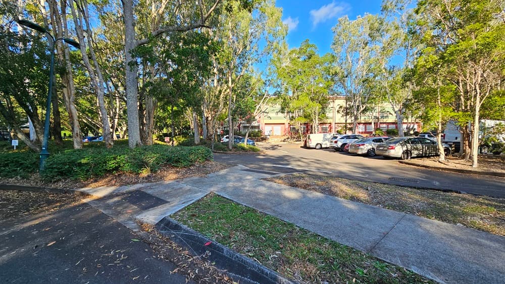 On the left, there is a pathway bordered by trees and bushes, with a lamp post visible among the foliage. On the right, there is a car park with several parked cars and more trees surrounding it. In the middle, a step free ramp and a concrete walkway leads from the foreground towards the background, intersecting with the car park. The background features a building partially obscured by trees. The floor consists of asphalt on the left and concrete in the middle and right areas.