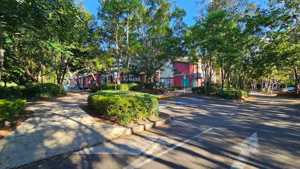 On the left, there is a concrete pathway bordered by green bushes and tall trees. On the right, there is an asphalt car park with white parking lines and more trees surrounding it. In the middle, a concrete walkway leads from the foreground towards the background, intersecting with the car park. The background features a building with red and white walls, partially obscured by trees.