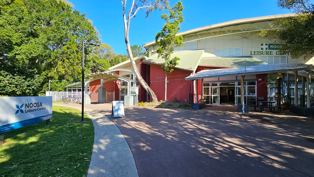 On the left, there is a sign for Noosa Leisure Centre next to a concrete pathway bordered by grass and trees. On the right, there is a large building with red and white walls, large windows, and a metal roof. The building has a covered entrance area with benches and tables. In the middle, the concrete pathway leads towards the building. The background features more trees and the continuation of the building. The floor is concrete, and the ceiling of the covered area is corrugated sheet.