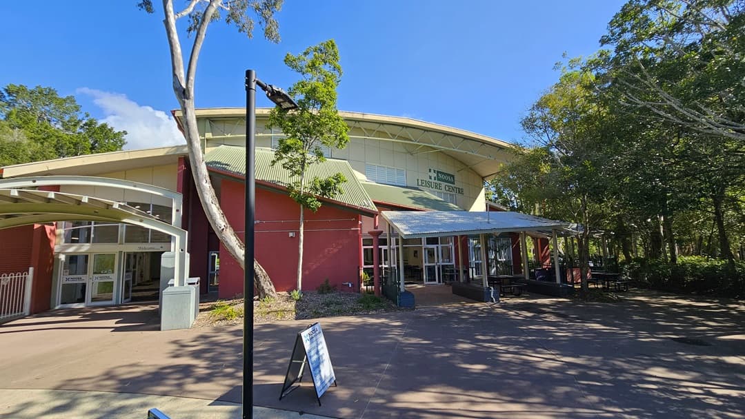A red and white building with large windows and a metal roof, featuring a covered entrance with a white metal fence. On the right, there is a continuation of the building with a sign reading "Noosa Leisure Centre" above the entrance. The entrance has a covered area with tables and chairs, surrounded by trees. In the middle, a concrete pathway leads to the entrance. The background includes more trees and greenery. The floor is concrete, and the ceiling of the covered area is metal.