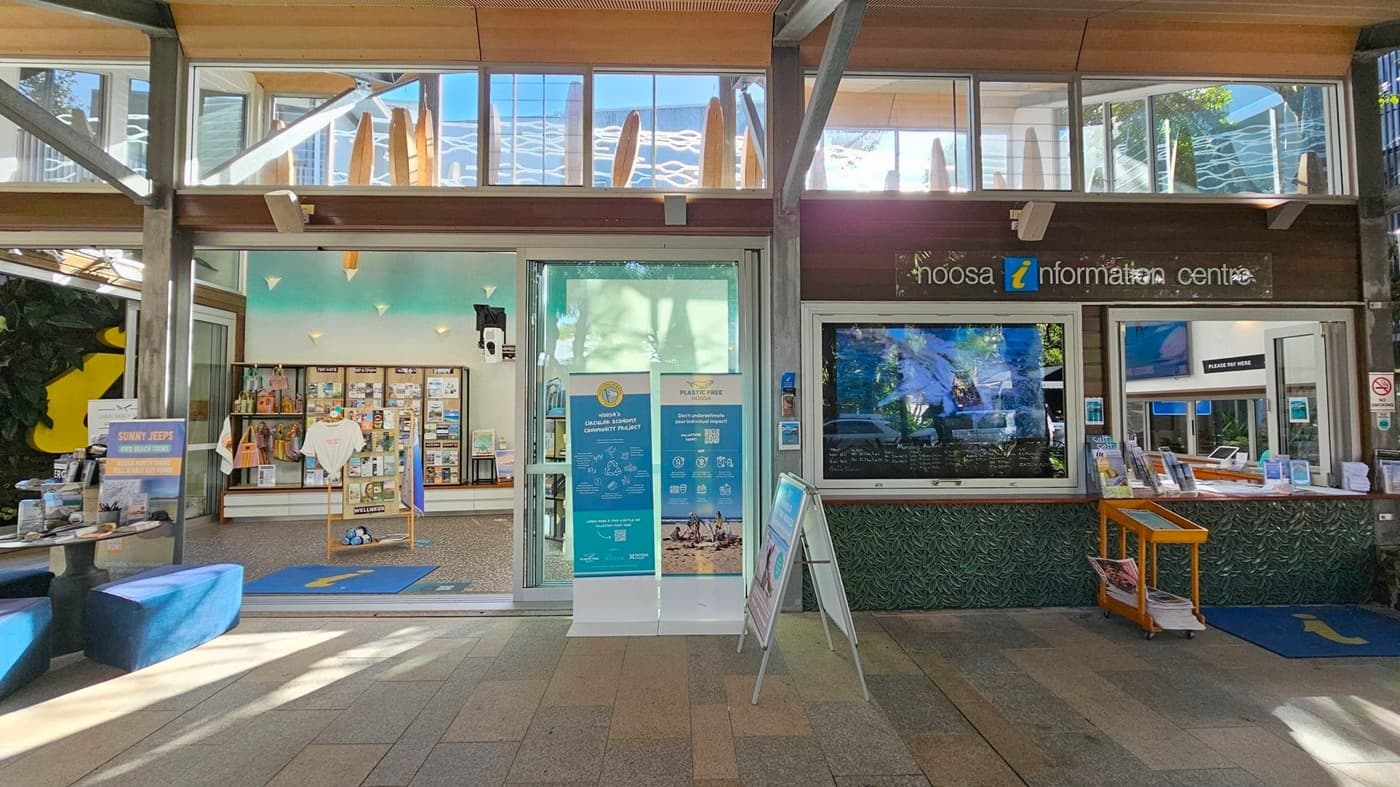 A visitor information centre with a modern design. On the left, there is a display area with brochures, a rack of T-shirts, and a sign for "Sunny Jeeps". In the centre, a glass door is flanked by posters about community projects and plastic-free initiatives. On the right, a service window with brochures and a digital screen. The ceiling has wooden panels, and the floor is tiled in various shades of green and brown. Surfboards are visible through the upper windows.
