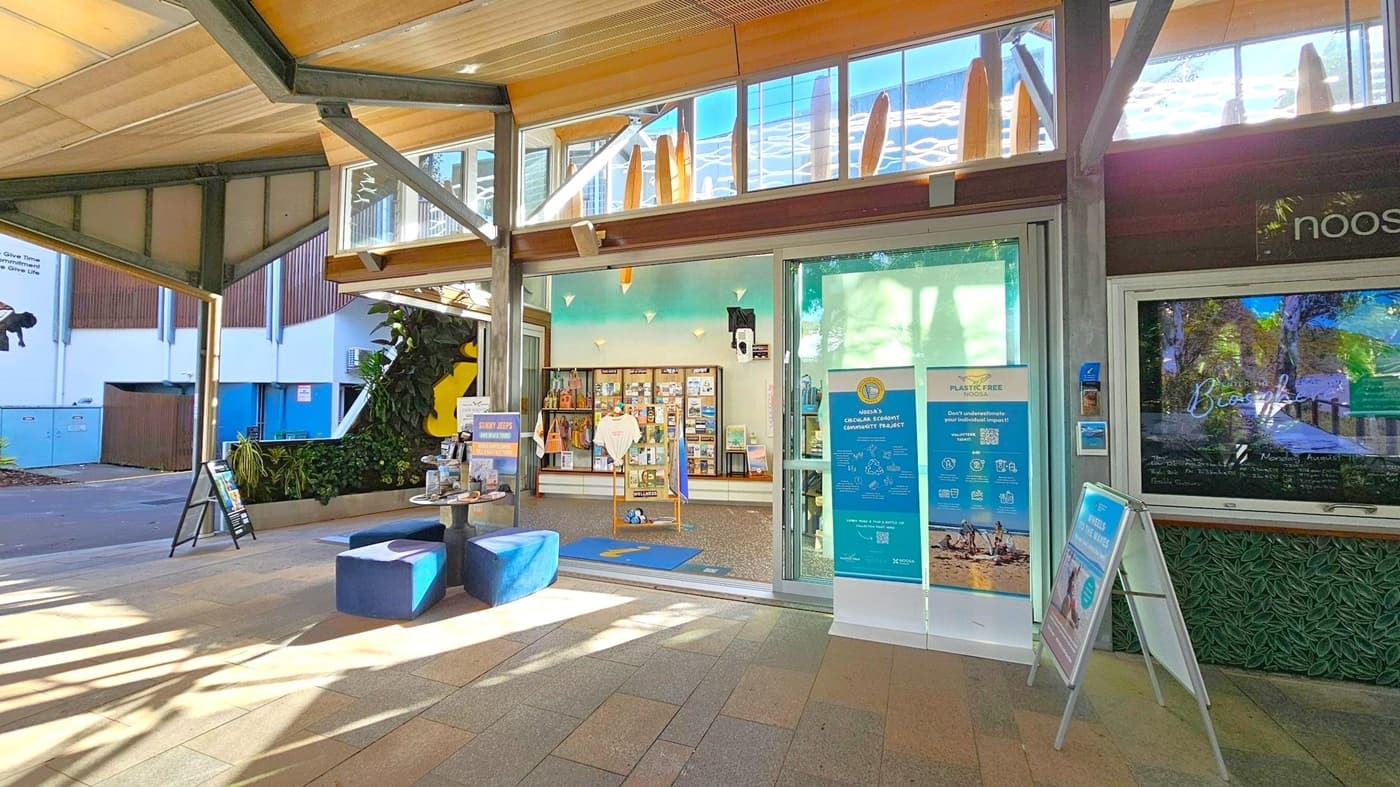 A modern indoor space with a high ceiling featuring exposed beams and large windows. In the centre, a shop entrance with glass doors displays various brochures and merchandise. To the left, a small seating area with blue cushioned stools and informational stands. The floor is tiled in a mix of earthy tones. On the right, a digital screen and a stand with promotional material. The background includes a wall with plants and a mural.