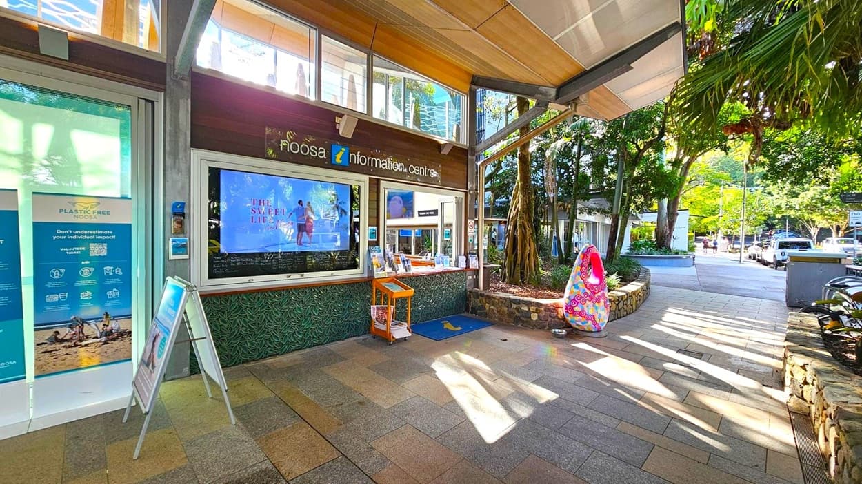 The image shows the entrance to the Noosa Information Centre. On the left, there are posters promoting a plastic-free initiative and a stand with brochures. In the centre, a large digital screen displays an advertisement. To the right, a colourful sculpture is placed near a stone planter with trees. The floor is paved with multicoloured tiles, and the ceiling has a wooden finish. Outside, a street with parked cars and trees is visible.