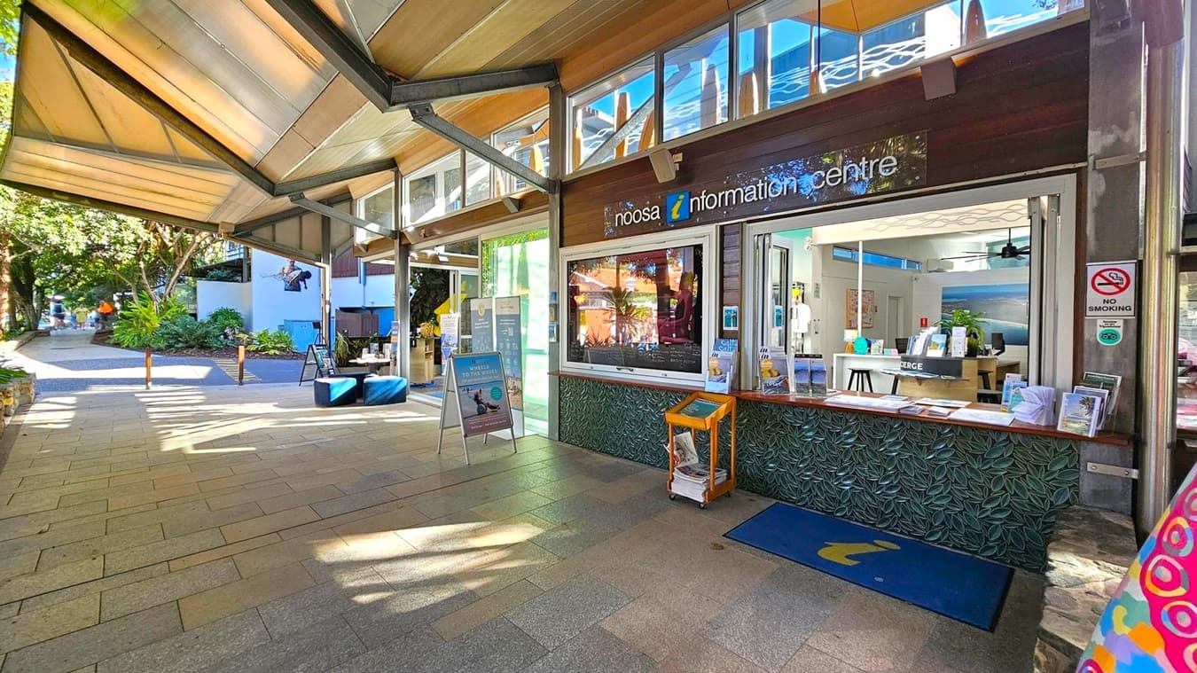 A covered walkway with a tiled floor leads to the Noosa Information Centre on the right. The centre has a large window displaying brochures and a "No Smoking" sign. Inside, there are informational materials and a counter. On the left, a path lined with greenery and trees extends into the distance. The ceiling is made of angled metal and glass panels, allowing natural light to filter through.