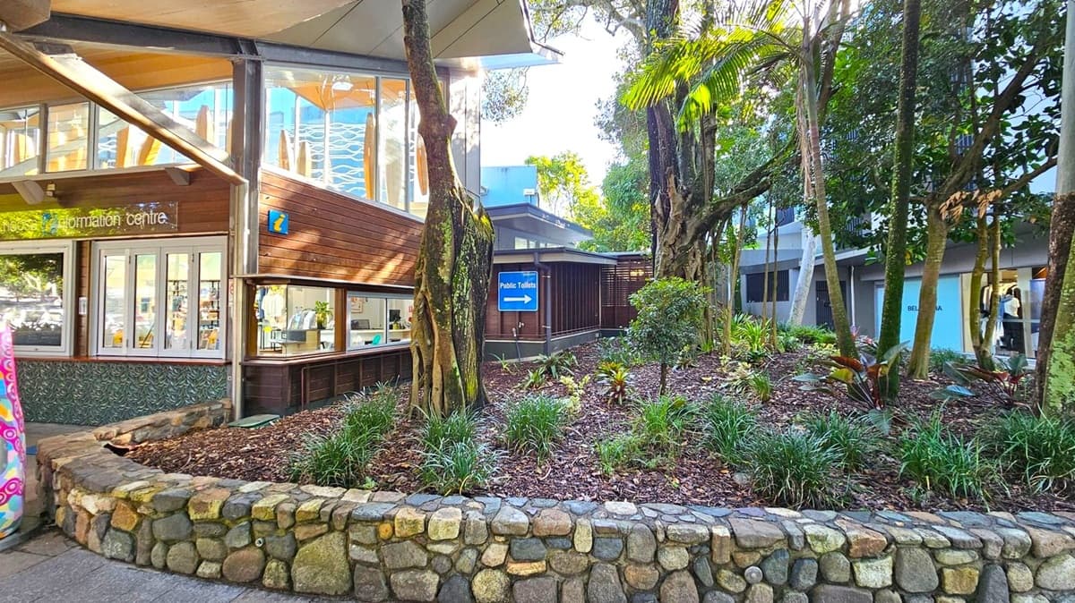 On the left, a building with a wooden exterior and large windows houses an information centre. A stone wall borders a landscaped area with trees and plants in the middle. To the right, a sign indicates the direction to public toilets. The background features more trees and a modern building with a grey facade. The ground is paved, and the ceiling of the building is visible with a canopy structure.