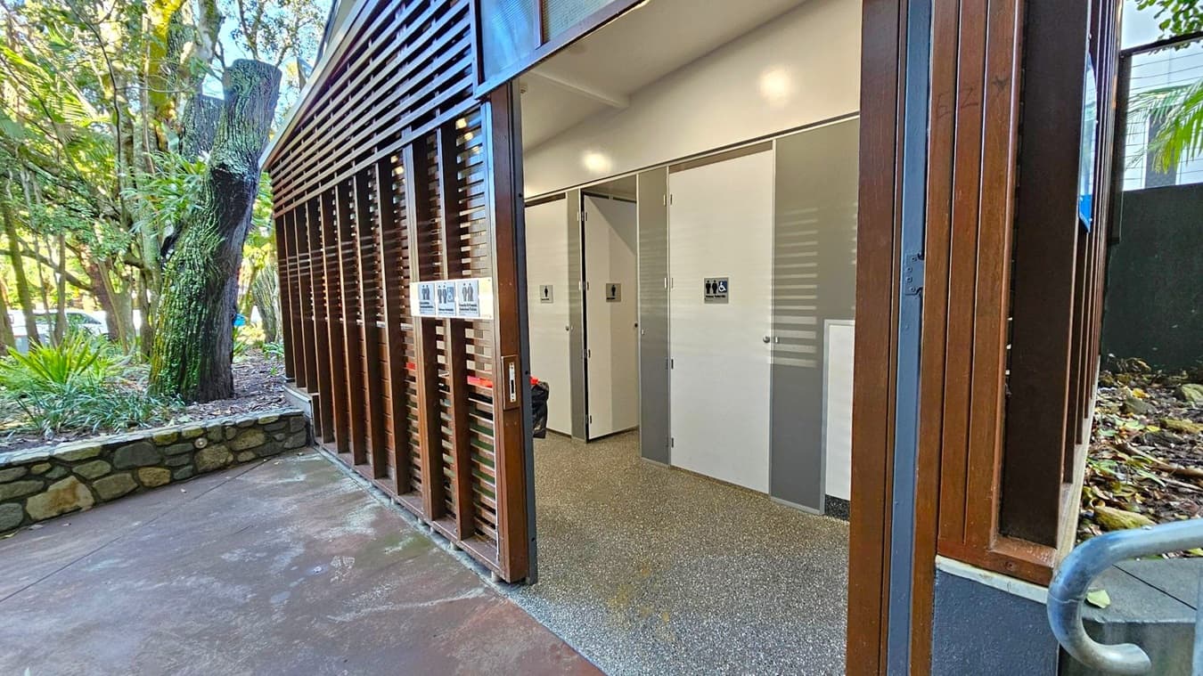 A public restroom facility with a modern design. On the right, there are two doors with signs indicating accessibility and gender-neutral use. The walls are light-coloured, and the floor is speckled grey. On the left, there is a wooden slatted structure with signage and a stone wall bordering a garden area. The garden features a large tree and various plants. The ceiling is plain and white, with recessed lighting.
