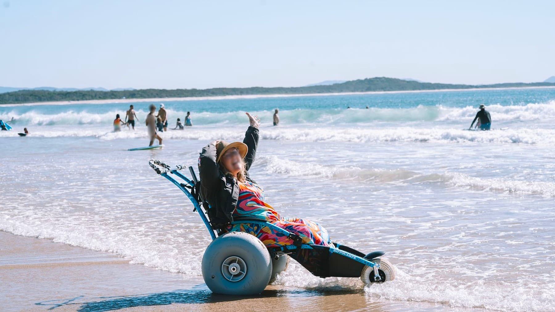 A person in a colourful outfit and hat is sitting in a beach wheelchair at the water's edge, with waves gently lapping around them. They have their arms raised, appearing relaxed. In the background, several people are enjoying the sea, some standing and others swimming. The shoreline curves gently, with distant land visible on the horizon. The sand is wet where the waves meet the beach.