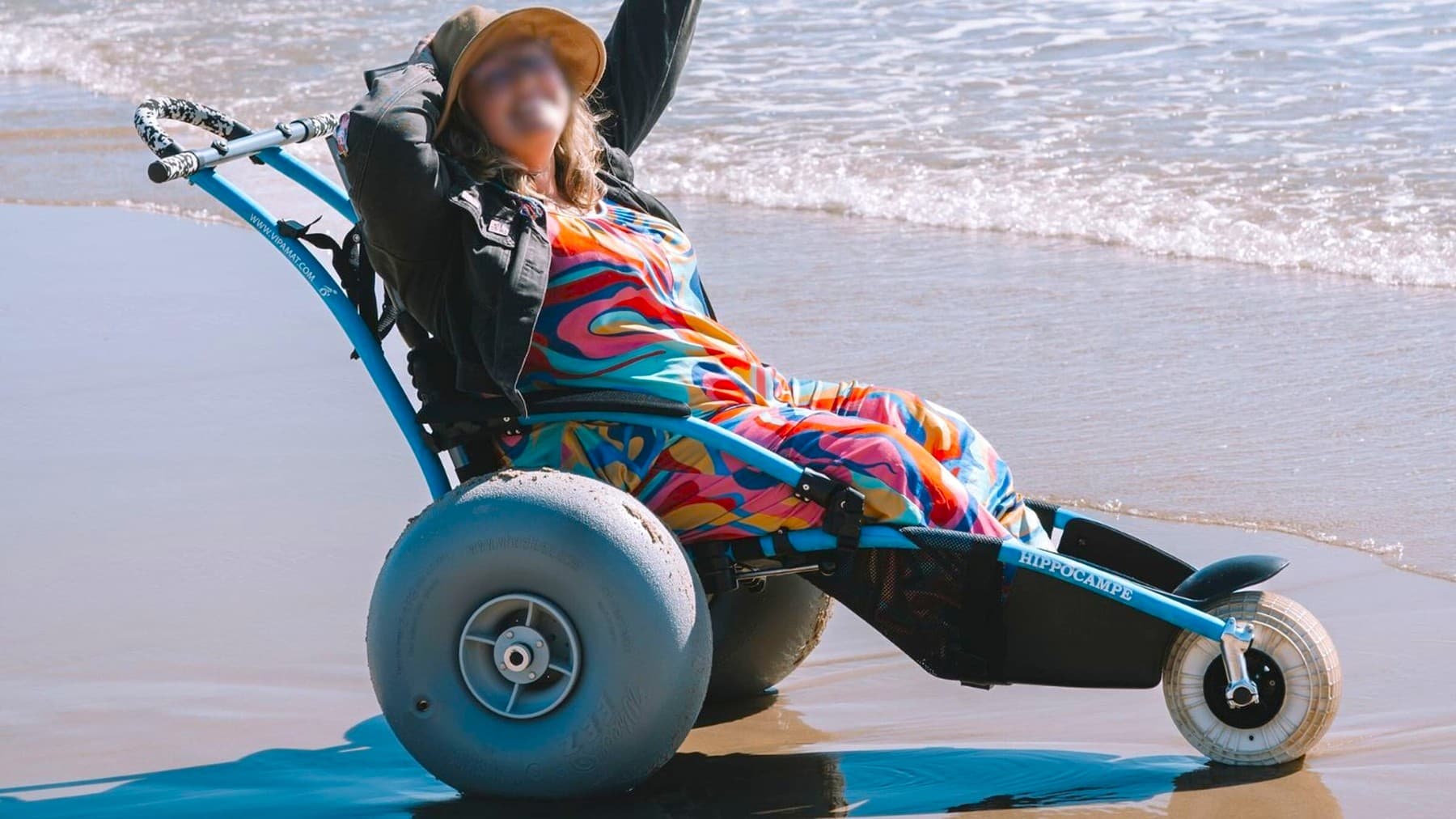 A person in a colourful, patterned outfit is sitting in a beach wheelchair with large, grey wheels designed for sand. They are wearing a hat and have one arm raised. The wheelchair is positioned on wet sand near the shoreline, with gentle waves in the background. The wheelchair has a blue frame with the word "Hippocampe" visible on the side.