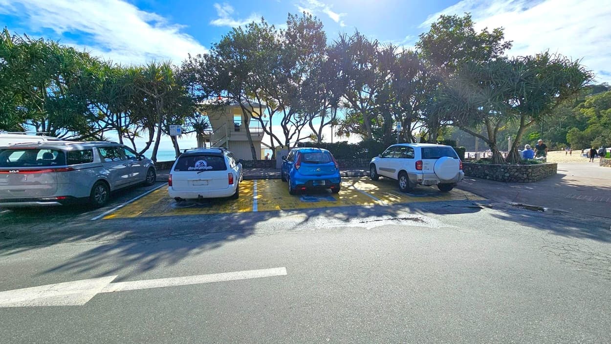 Four cars are parked in a row on a paved parking area. Behind the cars, a row of trees provides partial shade. A lifeguard tower is visible in the background, partially obscured by the trees. To the right, a few people are walking on a paved path. The ground is asphalt, and the sky is partly cloudy.