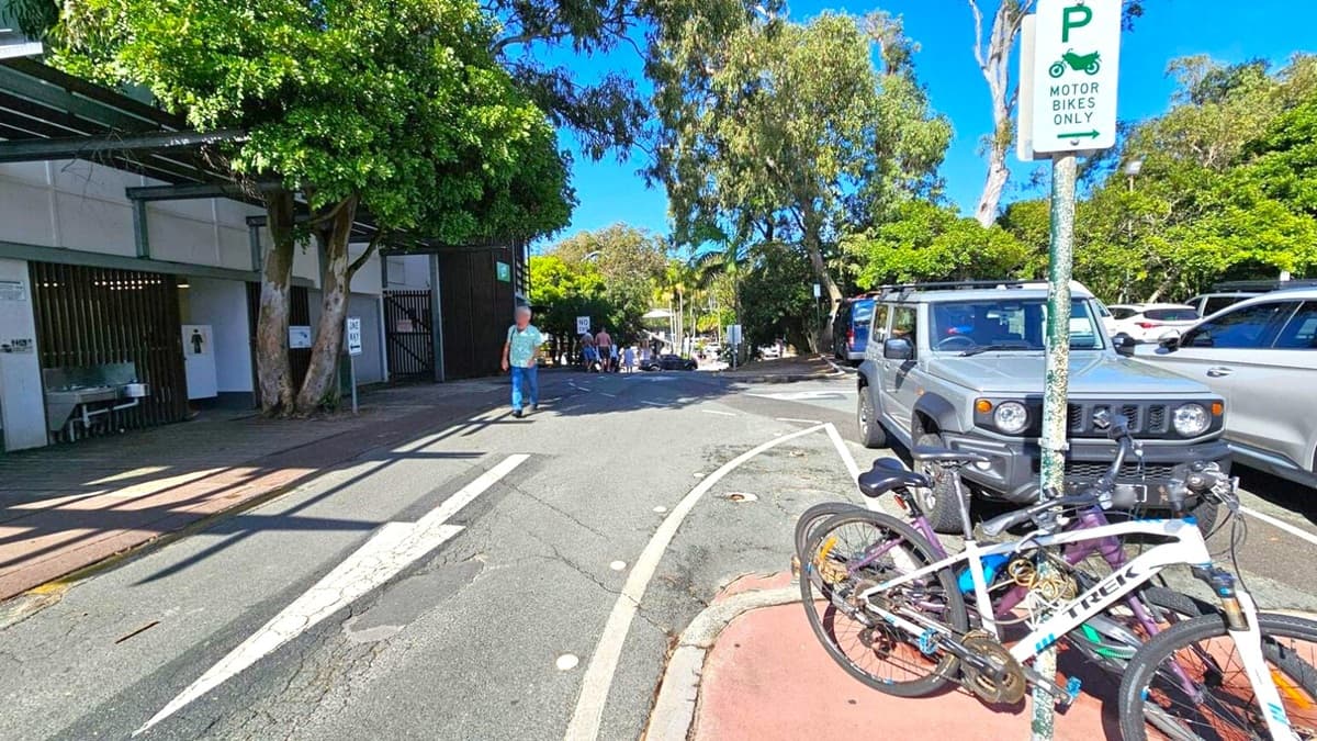 A street scene with a grey SUV parked on the right, next to a sign indicating "Motor Bikes Only". Two bicycles are locked to a pole in front of the SUV. On the left, a building with a public restroom is partially visible, with a tree growing close to it. A person walks along the pavement. In the background, more people are visible walking down the street, which is lined with trees and parked cars. The road has a "One Way" sign and a marked lane.