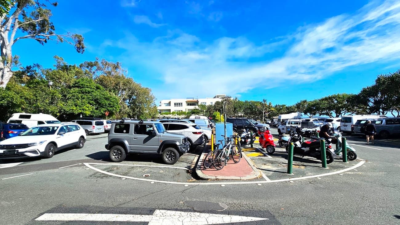 A busy car park with various vehicles, including a grey SUV and a white car, is in the foreground. Bicycles are parked on a small paved area in the centre, alongside several scooters. People are near the scooters, with one person sitting on a light blue scooter. In the background, there are trees and a light-coloured building. The ground is asphalt with visible markings.