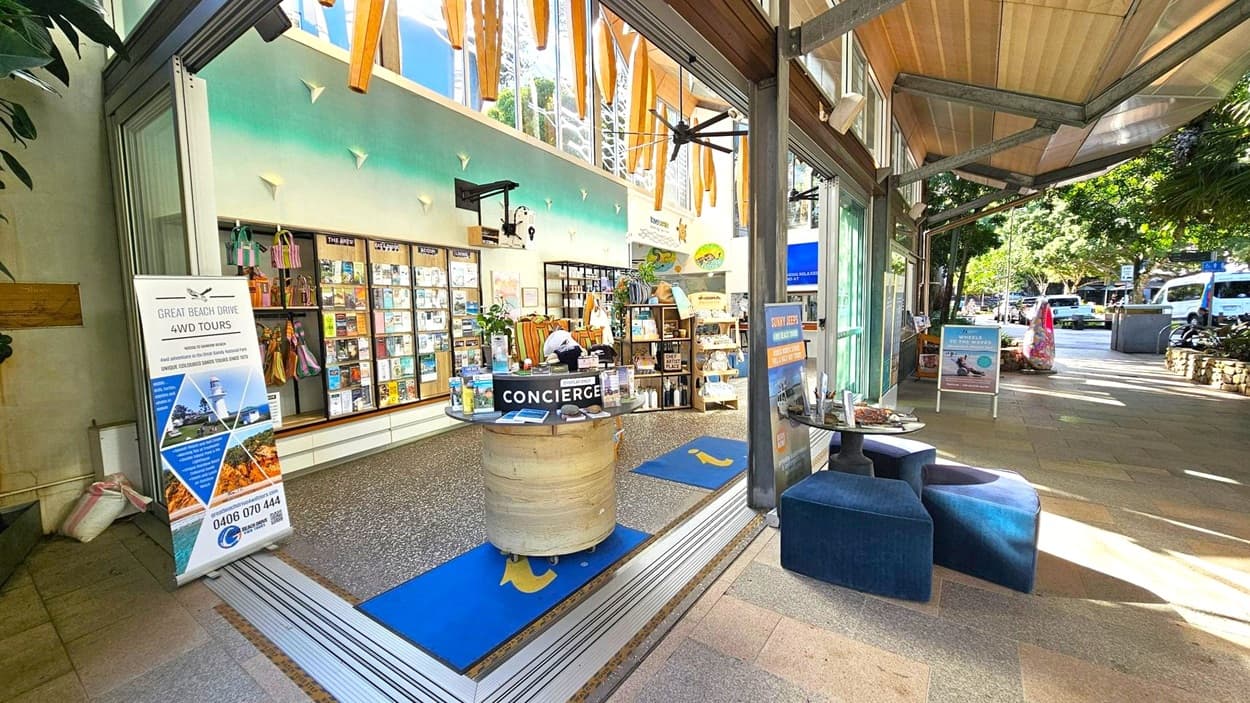 A tourist information centre with a concierge desk in the middle, featuring brochures and a sign. On the left, a display board with various pamphlets and a sign for 4WD tours. Shelves with merchandise are visible in the background. On the right, a small seating area with blue cushioned seats and a table. The exterior has a covered walkway with trees and parked vehicles in the distance. The ceiling has wooden beams and large windows with decorative elements.