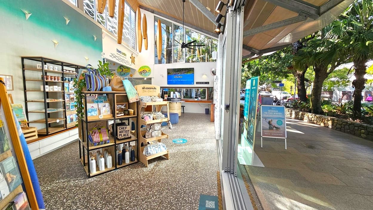Inside a shop with a terrazzo floor, shelves on the left display various products including bottles and bags. A central wooden display holds more items. The ceiling is wooden with large windows and surfboard decorations. A concierge desk is at the back. On the right, large glass doors open to a footpath lined with trees and a stone wall. Outside, signs and a view of the street with parked cars and pedestrians are visible.