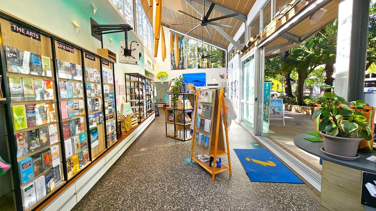 Interior of a visitor information centre with a terrazzo floor. On the left, a wall-mounted display holds brochures under categories like "The Arts" and "Eat & Drink". In the middle, a wooden stand and shelves display more brochures and books. The right side features large glass doors opening to a patio with trees visible outside. A potted plant sits on a counter near the entrance. The ceiling is high with exposed beams and large fans.