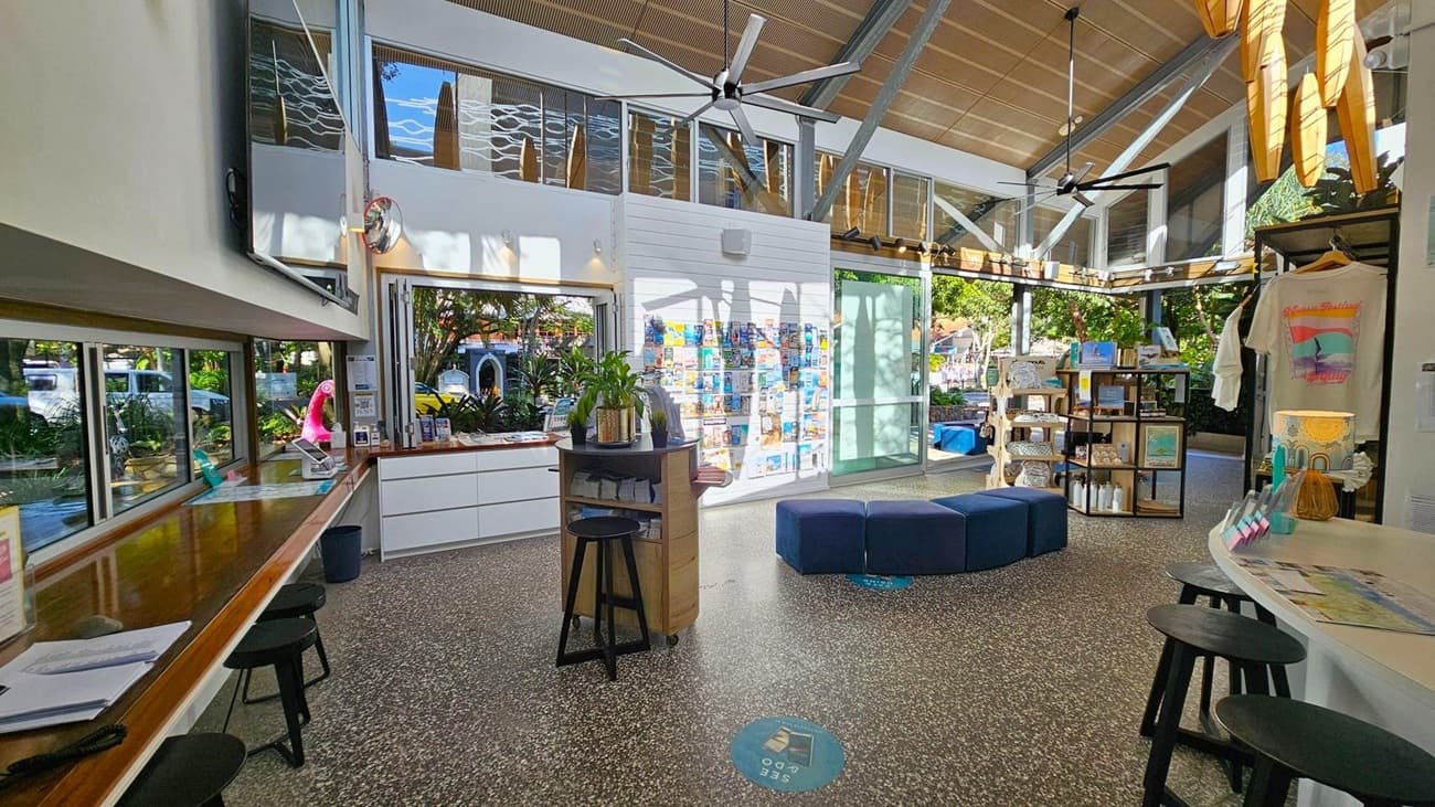 A spacious interior with a terrazzo floor and high, slanted wooden ceiling. On the left, a long wooden counter with stools and large windows overlooking greenery. In the centre, a display of brochures on a white wall and a round table with plants. To the right, shelves with merchandise, including a T-shirt and various items. Large ceiling fans are visible, and natural light streams through the windows, creating a bright atmosphere.