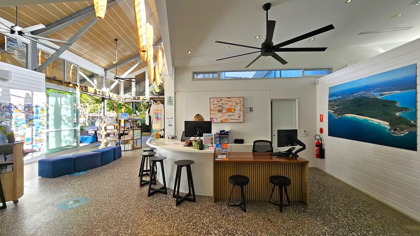 A spacious interior with a high, slanted ceiling featuring large wooden beams and ceiling fans. On the left, there are large windows and a glass door, allowing natural light to illuminate the room. A curved reception desk with black stools is in the centre, with a person working at a computer. On the right, a large aerial photograph of a coastal area is mounted on the wall. The floor is speckled, and there are shelves with merchandise near the windows.