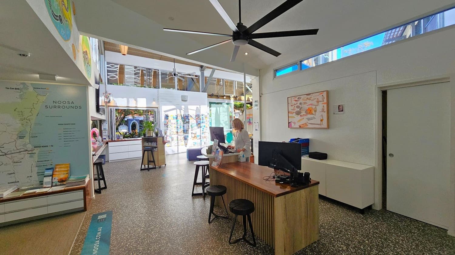 A modern interior with a large ceiling fan. On the left, a map labelled "Noosa Surrounds" is displayed on the wall above a low cabinet. In the centre, a person stands at a wooden counter with computers and stools. The right side features a white wall with a colourful artwork and a closed door. The floor is speckled, and the ceiling has narrow windows allowing natural light. In the background, there are brochures and a glimpse of an outdoor area.