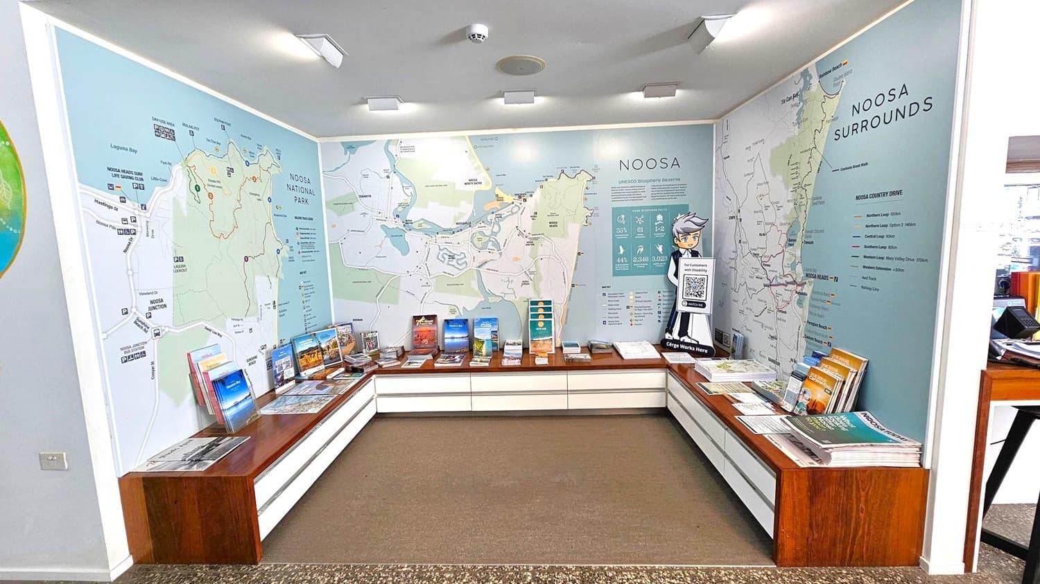 A room with large maps of Noosa National Park and its surroundings on the walls. Brochures and informational booklets are neatly arranged on wooden benches that line the walls. A cartoon character standee with a QR code is positioned on the right. The floor is carpeted, and the ceiling has several recessed lights.