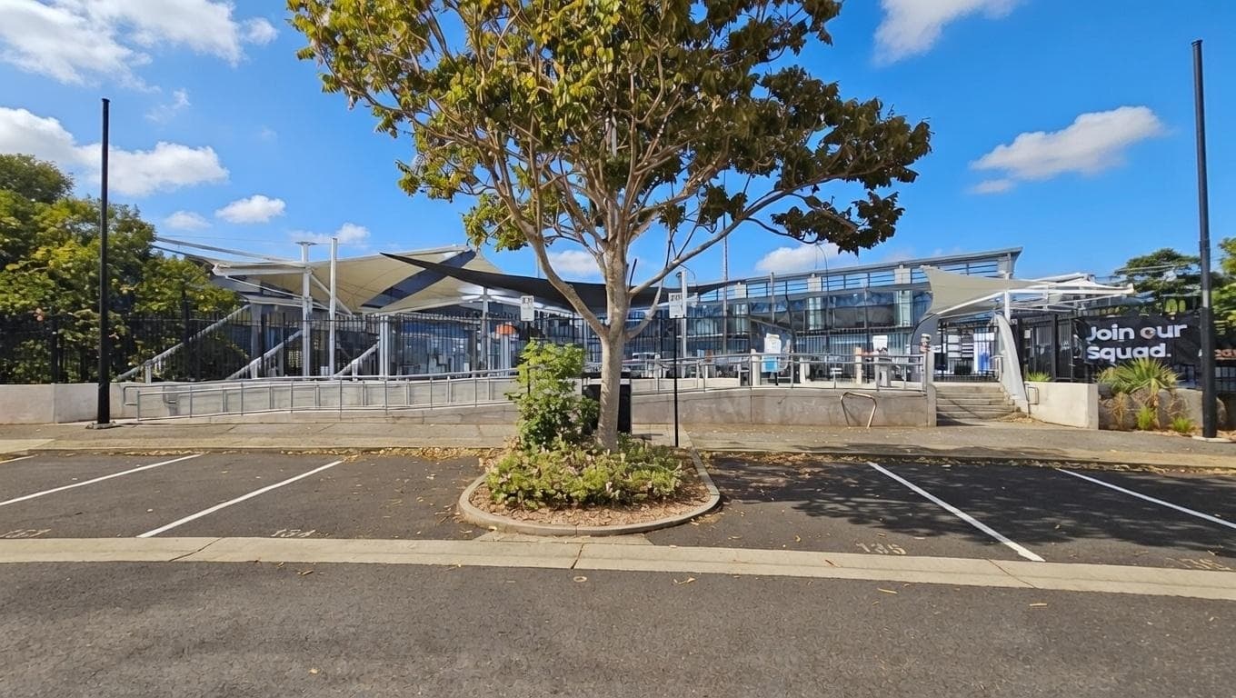 A modern building with a glass facade is in the background, featuring angular architectural elements. In the foreground, a tree with green leaves is centred in a small landscaped area surrounded by a car park. The car park has marked spaces and is mostly empty. On the right, a sign reads "Join our Squad." The area is well-lit, with tall streetlights visible.