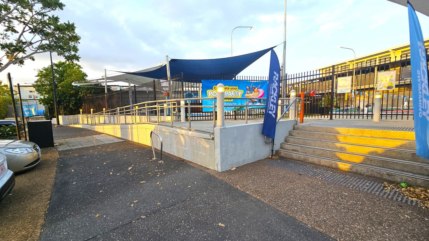 A paved pathway leads to a ramp and stairs on the right, bordered by a black metal fence. Blue banners with white text are positioned near the entrance. A colourful sign advertising a pool party is attached to the fence. A blue shade cloth is overhead. On the left, a parked car and a tree are visible. The ground is a mix of asphalt and concrete, with scattered leaves. A building is in the background.