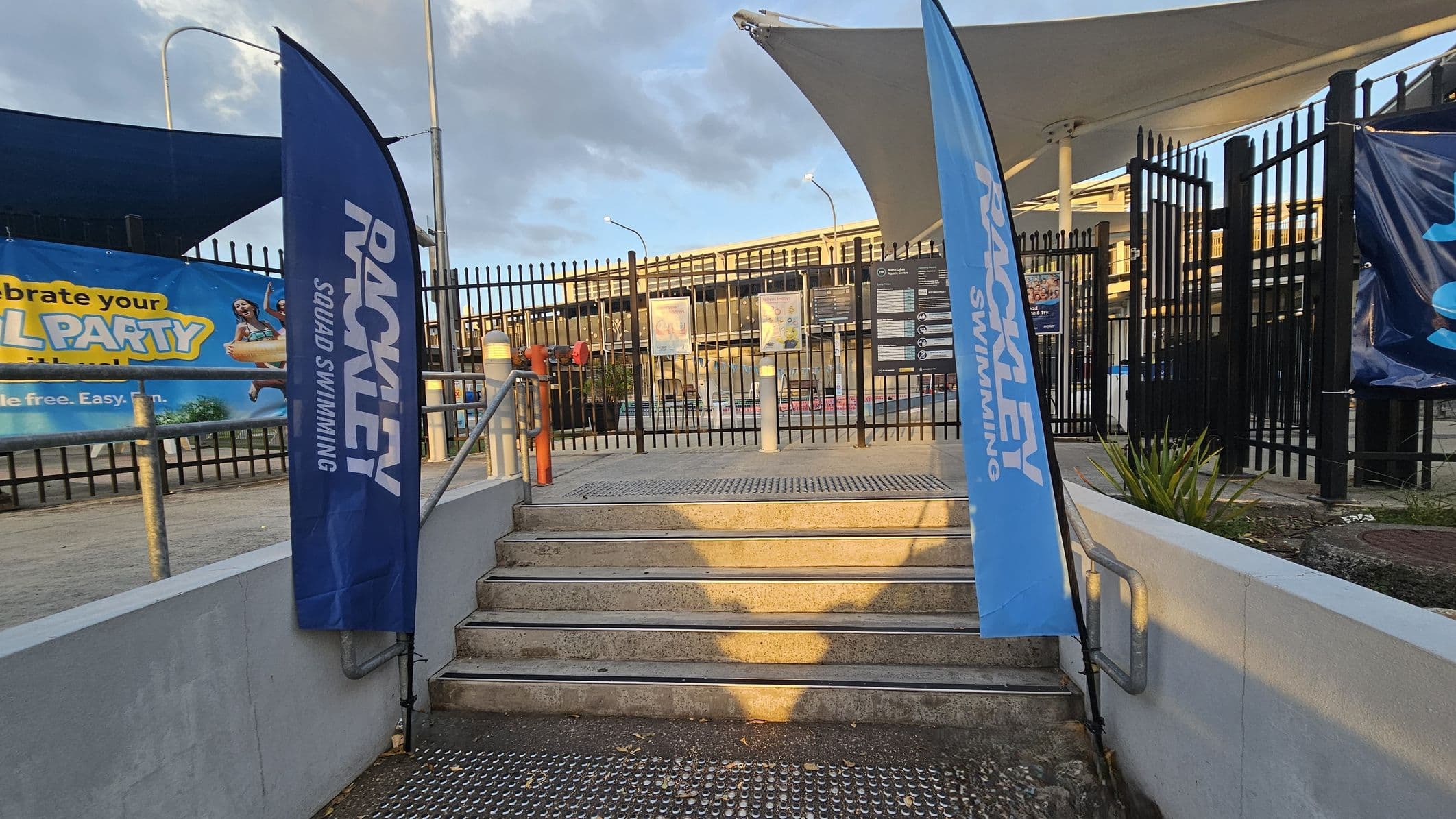 A set of concrete steps leads up to a gated entrance with black metal fencing. On either side of the steps are blue flags with "RACKLEY SQUAD SWIMMING" written on them. To the left, a colourful banner advertises a pool party, featuring an image of a person with a float. The ground is paved, and there are small plants near the entrance. Above, a large white canopy provides shade. The area is well-lit with street lamps.