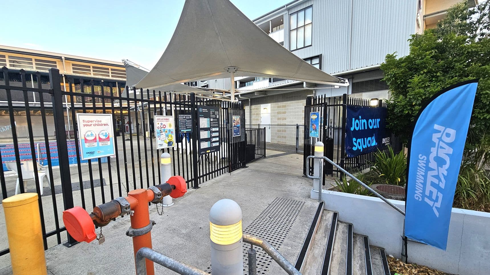 A swimming pool entrance with a black metal fence and gate. On the left, there are signs about supervising children and joining a swimming squad. A large white shade structure is above the entrance. On the right, a blue flag with "RACKLEY SWIMMING" and a banner saying "Join our Squad" are visible. Steps with metal railings lead up to the entrance. A building with large windows is in the background, and there are plants near the entrance.