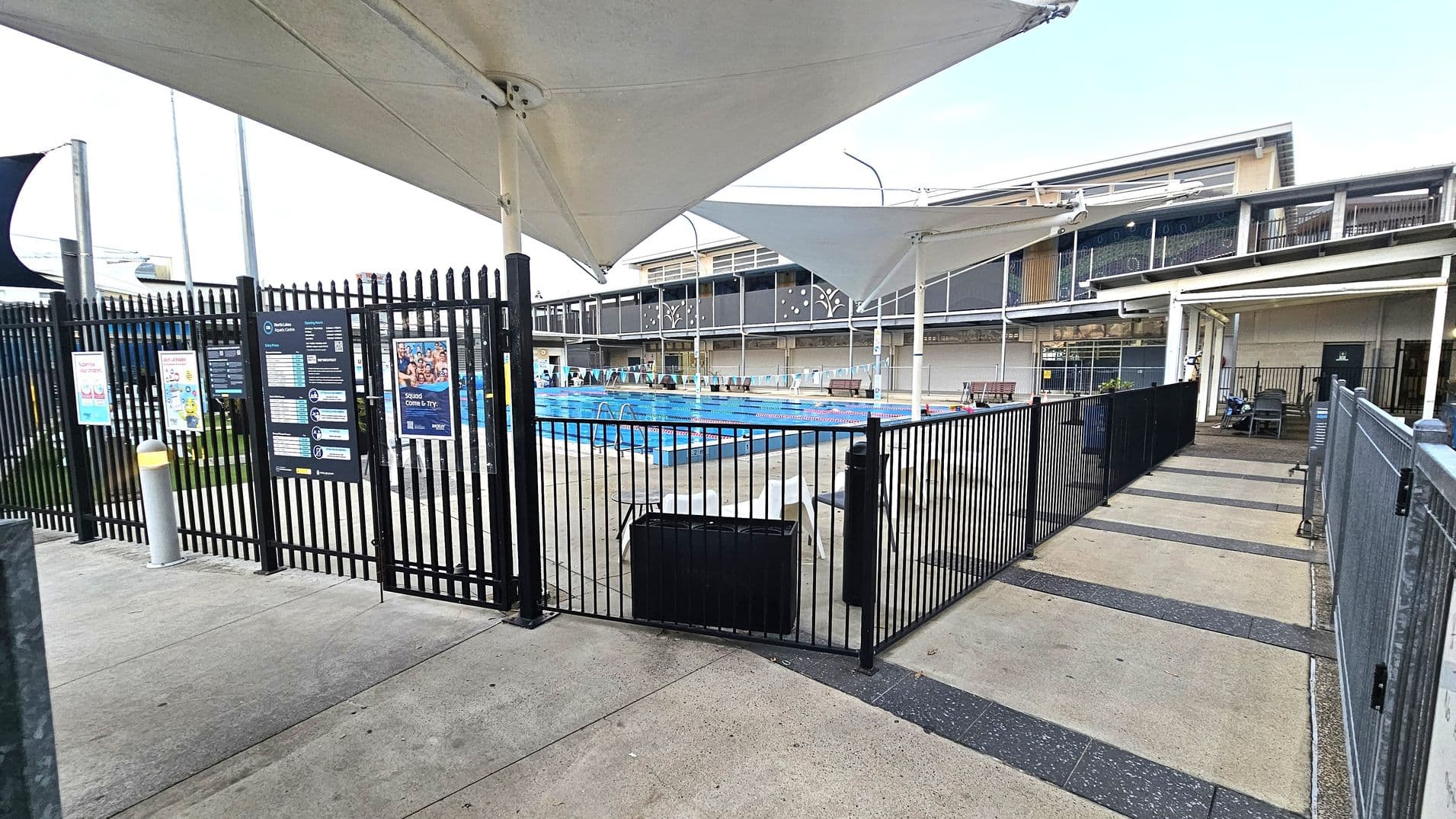 A swimming pool area is visible behind a black metal fence with informational signs and posters. The pool is surrounded by a paved area with white chairs and shaded by large triangular canopies. In the background, there is a building with a modern design featuring large windows and decorative panels. The foreground shows a concrete path leading to the pool entrance, with a bollard and a bin nearby.