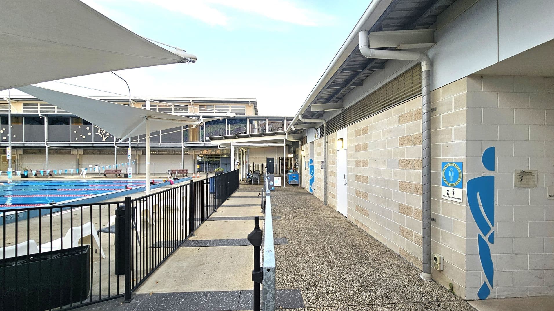 On the left, there's a swimming pool with lane dividers and a black metal fence. White shade sails are above the pool area. On the right, a building with light-coloured brick walls features a blue figure symbol near a door, indicating a toilet or changing area. The ground is a mix of concrete and gravel. In the background, there are benches and a building with large windows.