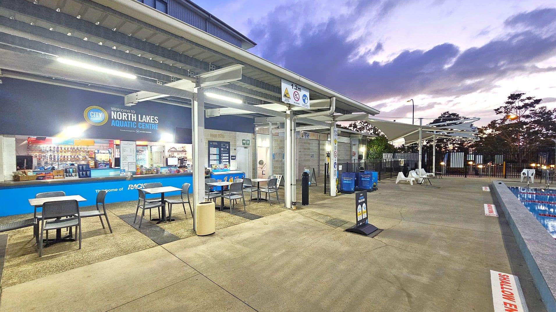Outdoor area of an aquatic centre with a covered seating area on the left, featuring tables and chairs. A sign reads "Welcome to North Lakes Aquatic Centre" above a counter displaying snacks and drinks. The right side shows a pool with a "Shallow End" marking, surrounded by a fence. White sun loungers and umbrellas are near the pool. The ground is a mix of concrete and textured surfaces, and the ceiling is a metal structure with lights. Trees and a cloudy sky are visible in the background.