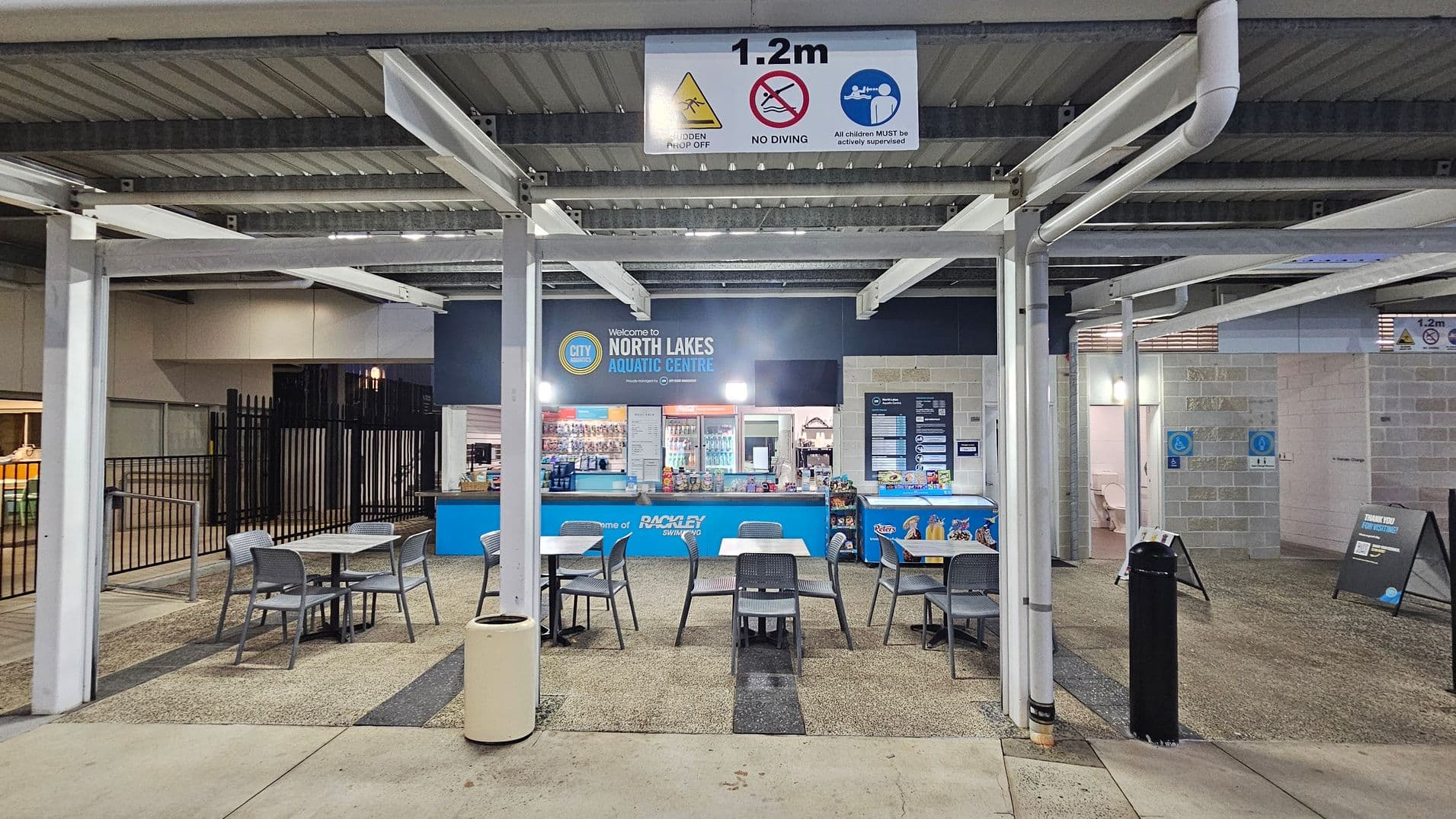 A covered outdoor seating area with several tables and chairs on a textured concrete floor. In the background, a counter with a blue sign reads "Welcome to North Lakes Aquatic Centre." Behind the counter, shelves display various snacks and drinks. A sign above warns of a "1.2m hidden drop off" and "no diving." The ceiling is metal with exposed beams. To the right, there are signs indicating accessible facilities.